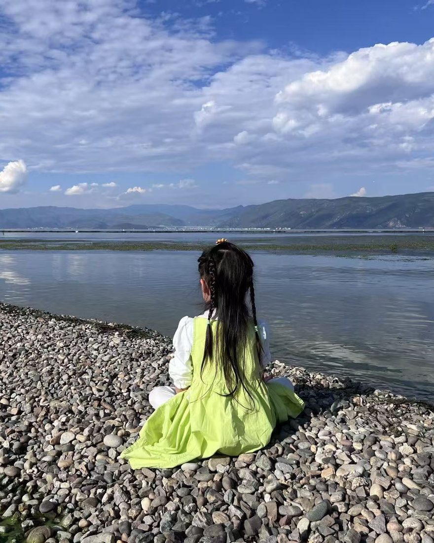 A child sitting on a pebble shore, facing the mountains across Erhai Lake in Dali, China / 女儿坐在洱海边的卵石滩上，望着远处的苍山